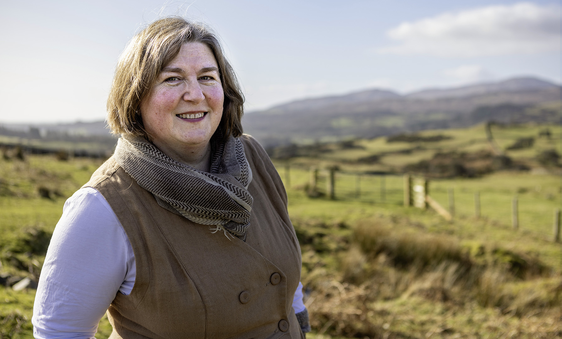Woman smiles at camera. She is white, with short brown hair framing her face. In the background are lush green hillsides. She is wearing soft natural colours, of light honey-ed brown.